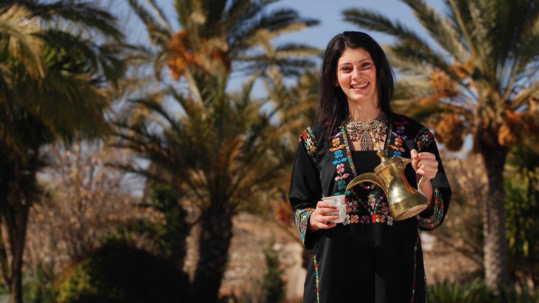 Jordaniesche Frau in traditioneller Kleidung schenkt Tee aus Kanne ein.