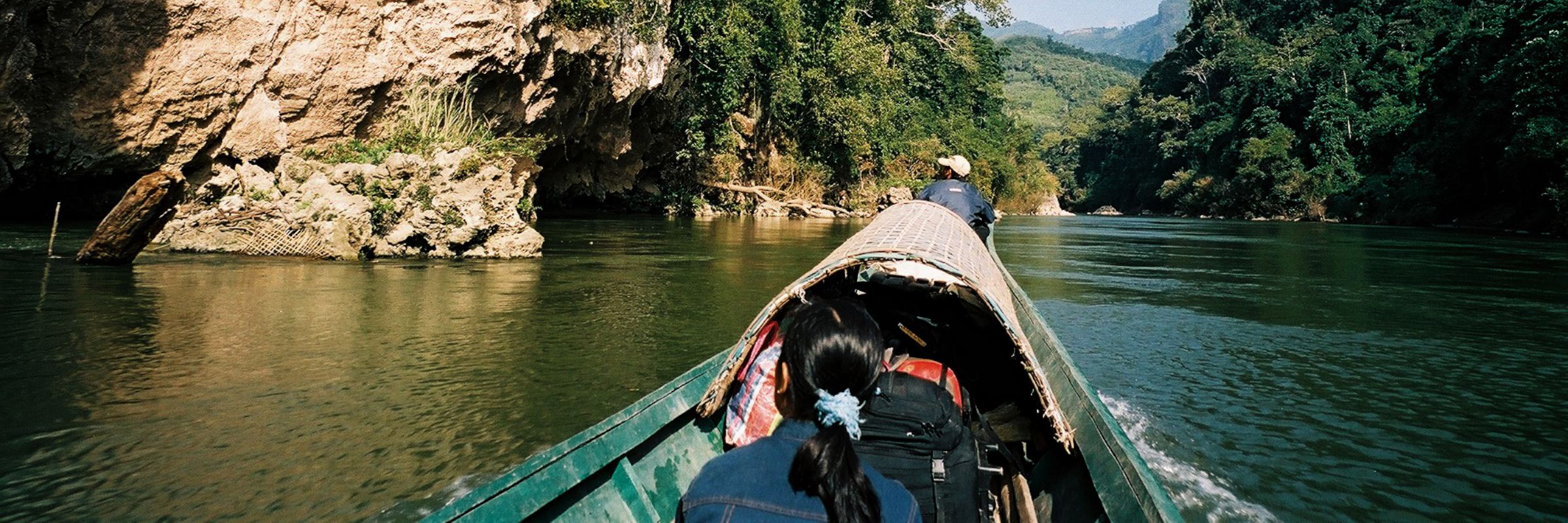 Zwei Einheimische fahren mit einem landestypischen Boot durch eine idyllische Flusslandschaft im Norden von Laos.